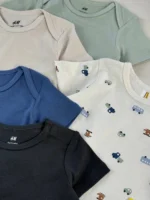 نمای نزدیک بادی آستین کوتاه نوزادی پسرانه H&M در رنگ‌های کرم، آبی، مشکی و سفید با طرح وسایل نقلیه همراه با لوگوی پنبه ارگانیک
